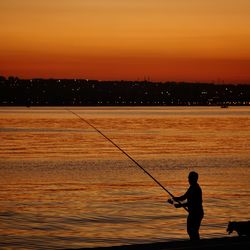 Silhouette mature man fishing at beach against sky during sunset