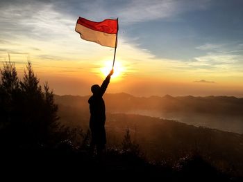 Man holding flag while standing on mountain against sky during sunset