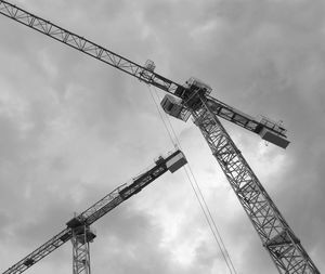 Low angle view of cranes at construction site against cloudy sky