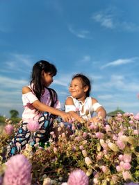 Cute sisters crouching by flowering plants against sky