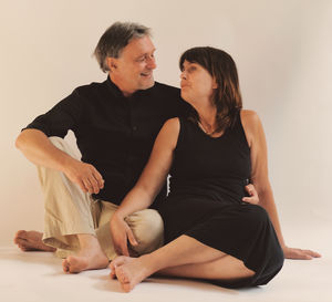 Couple in black dresses sitting on the floor against beige background smiling at each other