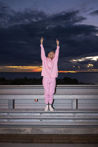 Young woman with pink hair wearing pink track suit shirt standing on road barrier at dusk