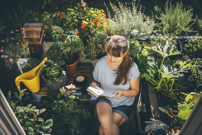 Woman sitting and reading book near plants in balcony