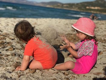 Side view of girl throwing sand on baby boy at beach during sunny day