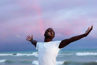 Happy man with arms outstretched standing at beach against sky during sunset