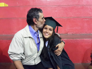 Father kissing graduate daughter while sitting against red wall