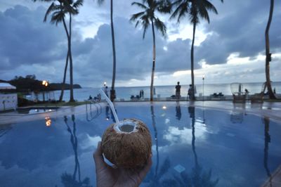 Cropped hand holding coconut water by swimming pool near sea against cloudy sky