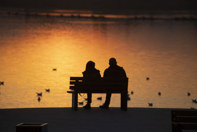 Silhouette couple sitting on bench by sea against sky during sunset
