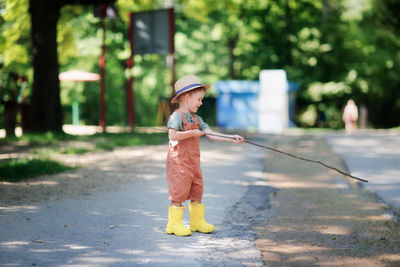 Cute european boy in hat and overalls in summer with stick and fishing rod plays with stream on road 