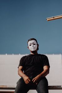 Young man wearing mask while siting on railing against clear sky against 