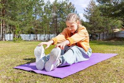 A teenage girl goes in for sports in nature, on a sunny summer day sits on a sports mat, stretches. 