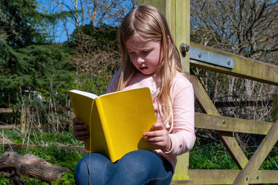 Girl enjoying reading from yellow textbook while sitting on wooden fence.