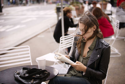 Woman in jacket reading book while sitting at sidewalk cafe