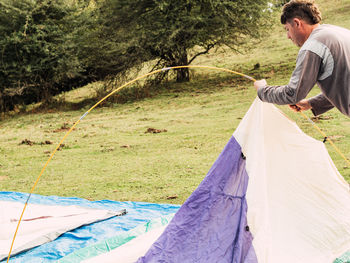 Side view of young male traveler in casual clothes opening camping tent on green grassy meadow during trip in countryside