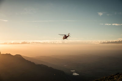 Helicopter flying over mountains against sky during sunset