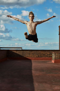 Masterful young blond ballet dancer in black leotard performing exciting jump in splits raising hands up on roof of building on sky background in sunny day