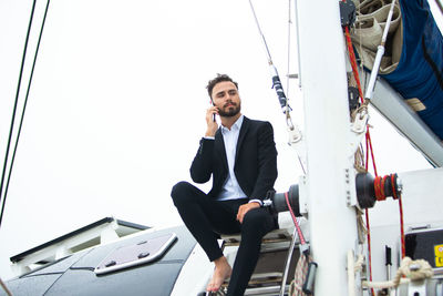 Low angle view of businessman talking on phone while sitting on boat against clear sky