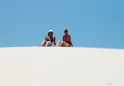 Friends sitting on sand dune against clear blue sky during sunny day