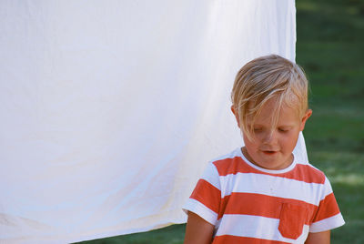 Thoughtful boy standing against white textile hanging outdoors