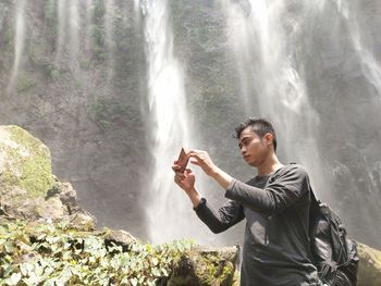 Young man photographing from mobile phone against waterfall