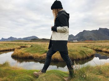 Side view of woman walking by pond amidst grassy field against cloudy sky