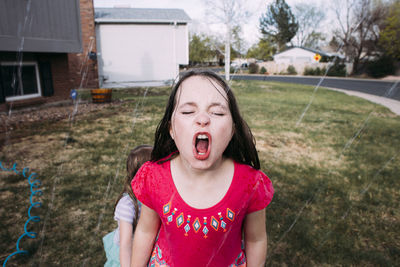 High angle view of playful girl enjoying in sprinkler with sister in background at yard