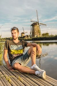 Portrait of man sitting on jetty over lake against traditional windmill