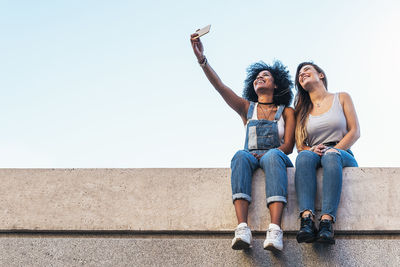 Low angle view of female friends taking selfie while sitting on retaining wall