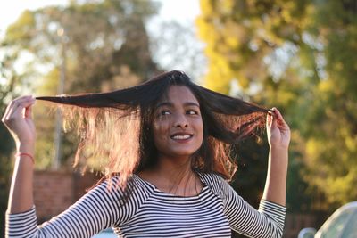 Smiling young woman playing with hair while standing against trees