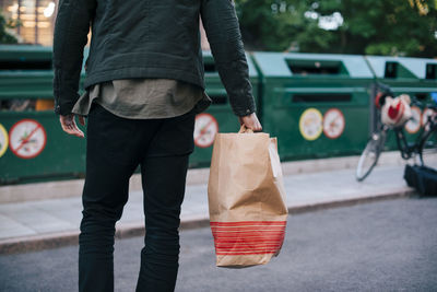 Midsection of man carrying paper bag against garbage cans