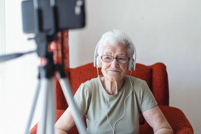Content senior lady in headphones sitting in cozy armchair at home and enjoying songs while looking away
