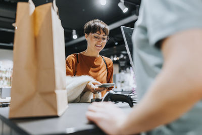 Smiling mature woman making payment through smart phone at checkout counter in electronics store