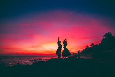 Silhouette women posing at shore against sky during sunset