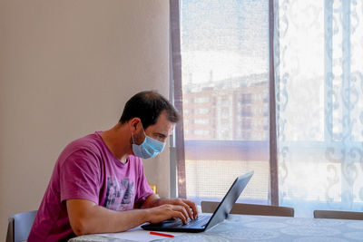 Side view of man wearing flu mask using laptop at home