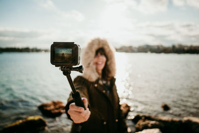 Woman taking selfie with camera while standing against lake