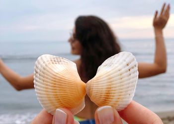 Cropped hands holding seashells against woman standing at beach
