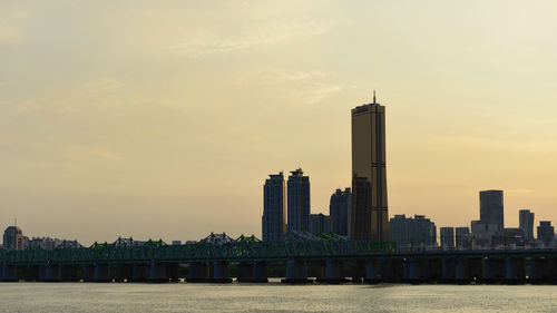 Modern buildings by river against sky during sunset