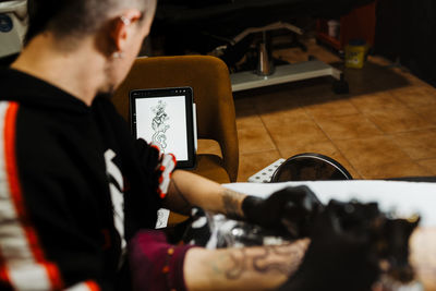 From above cropped unrecognizable man with using tattoo machine to make tattoo on leg of crop customer during work in salon