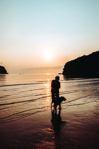 Silhouette senior woman with dog standing at beach against sky during sunset