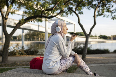 Positive arab female in hijab sitting on stone staircase near backpack and using cellphone after outdoor fitness training and listenig music on headphones