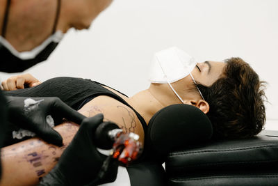 Crop male tattooist with machine making tattoo on arm of woman lying on table with closed eyes in salon