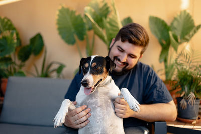 Young man sitting and smiling holding his dog while playing with paws. the dog is looking at camera