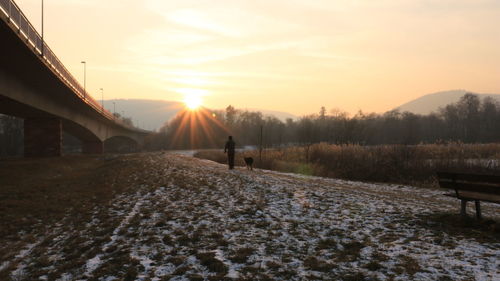 Rear view of person walking with dog on snow covered field during sunset