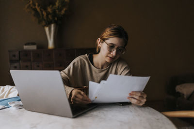 Non-binary person reviewing bills by laptop on table at home