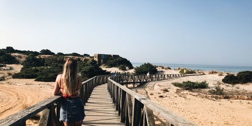 Rear view of woman standing on boardwalk by sea against clear sky