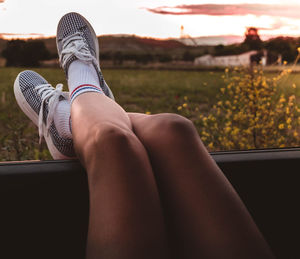 Low section of woman relaxing on car window against sky during sunset