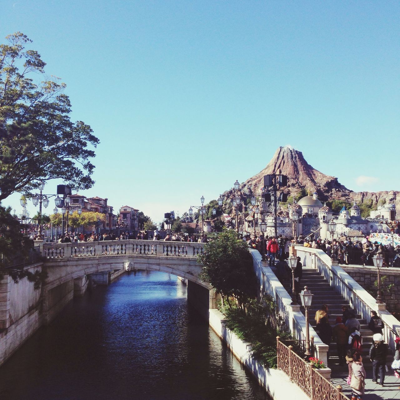 architecture, built structure, clear sky, building exterior, water, copy space, tree, blue, canal, river, bridge - man made structure, large group of people, connection, city, railing, incidental people, bridge, day, waterfront, mountain