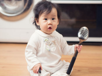 Cute baby girl sitting with ladle on hardwood floor at home