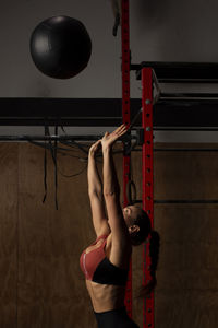 Side view of concentrated young fit sportswoman with long dark hair in activewear throwing heavy medicine ball and looking up during training in gym