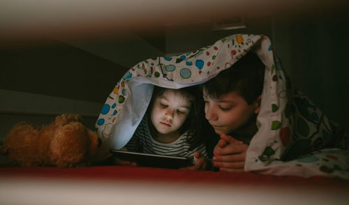 Siblings using digital tablet under blanket at home